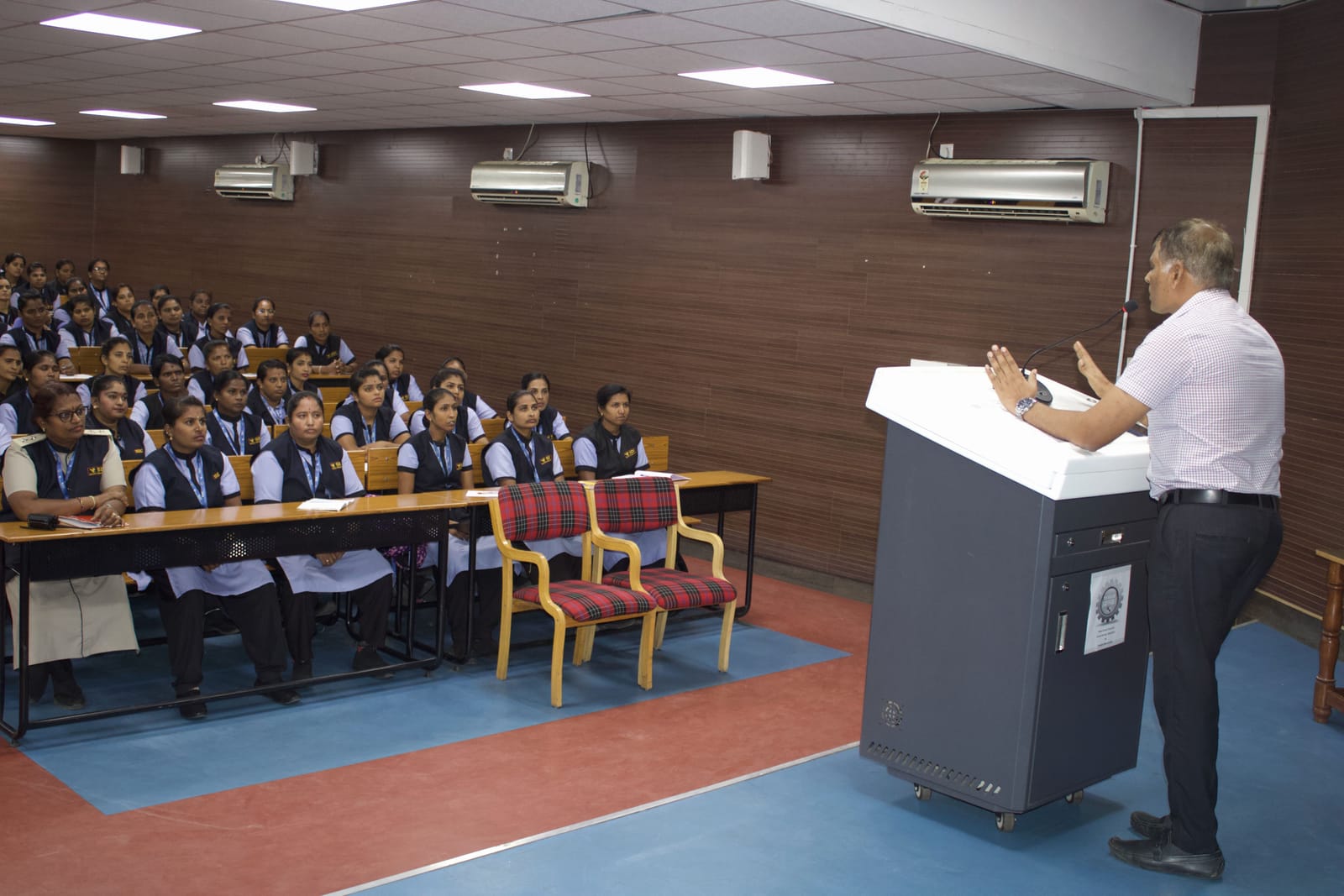 A trainer at the lectern addressing a hall of SDC guards and lady guards